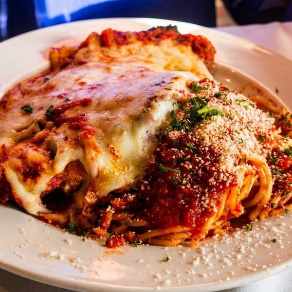 A plate of spaghetti with red sauce, melted cheese, and parmesan, with a person's hands nearby.