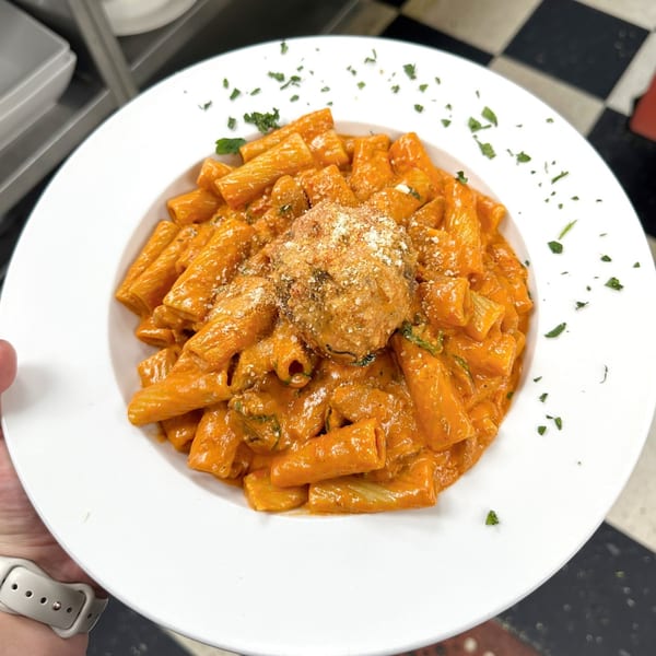 Rigatoni pasta in a creamy red sauce with a large meatball, garnished with parsley and parmesan.