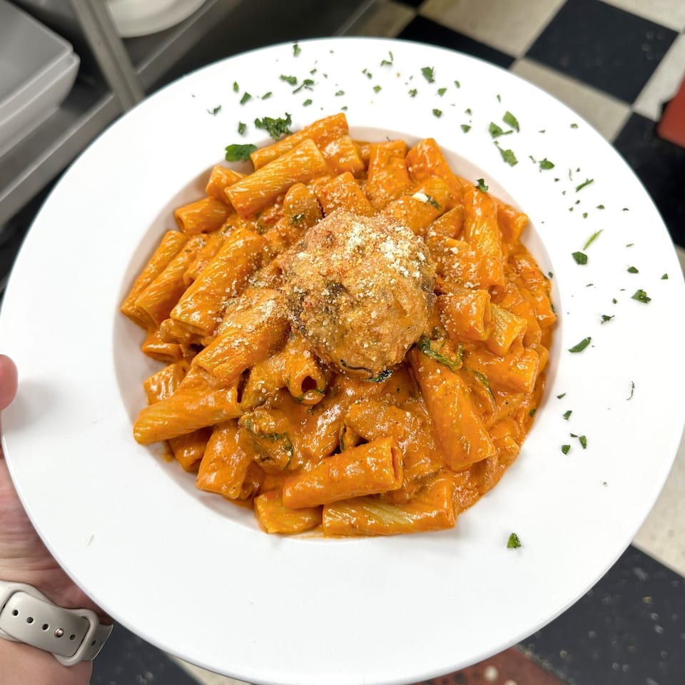 Rigatoni pasta in a creamy red sauce with a large meatball, garnished with parsley and parmesan.