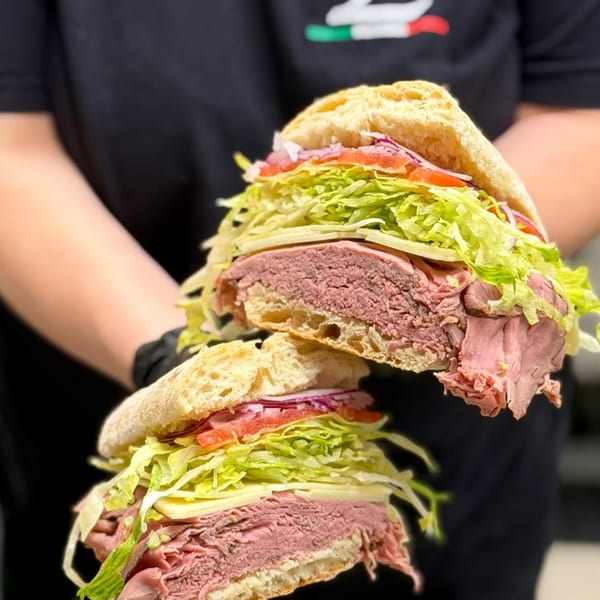 A person in a black shirt holding a large sandwich cut in half, revealing roast beef, cheese, lettuce, and tomato.