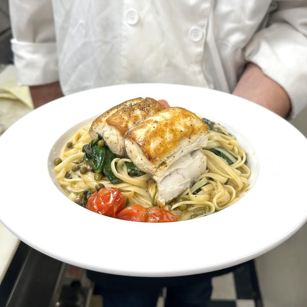 Chef holding a plate of pan-seared fish over linguine pasta with spinach, tomatoes, and capers.