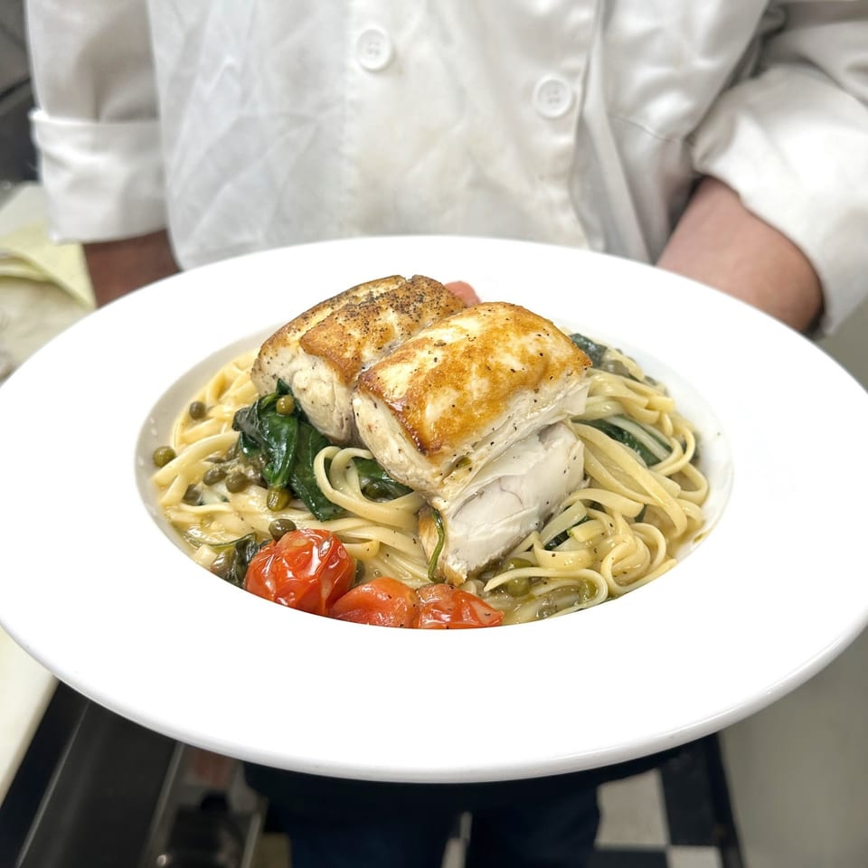 Chef holding a plate of pan-seared fish over linguine pasta with spinach, tomatoes, and capers.