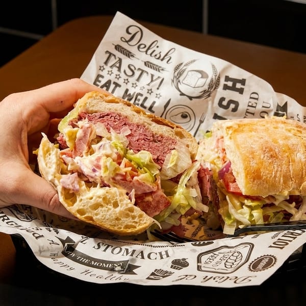 A hand holds a large deli sandwich with corned beef, coleslaw, and lettuce on ciabatta bread.