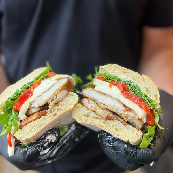 Gloved hands holding a sliced chicken cutlet sandwich with mozzarella, roasted peppers, and arugula.